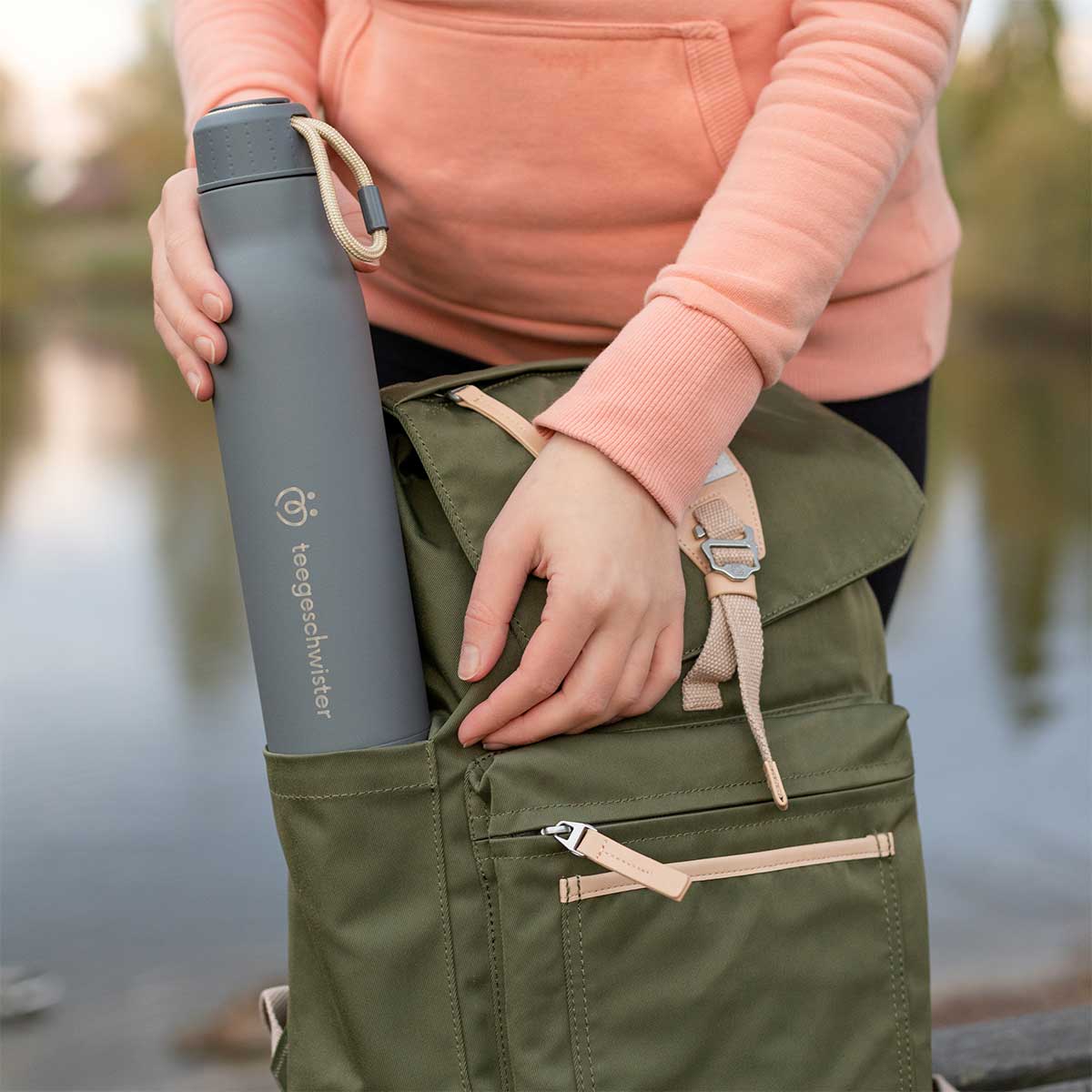 Das Bild zeigt wie eine graue Edelstahl Thermosflasche in einen grünen Rucksack gesteckt wird.
