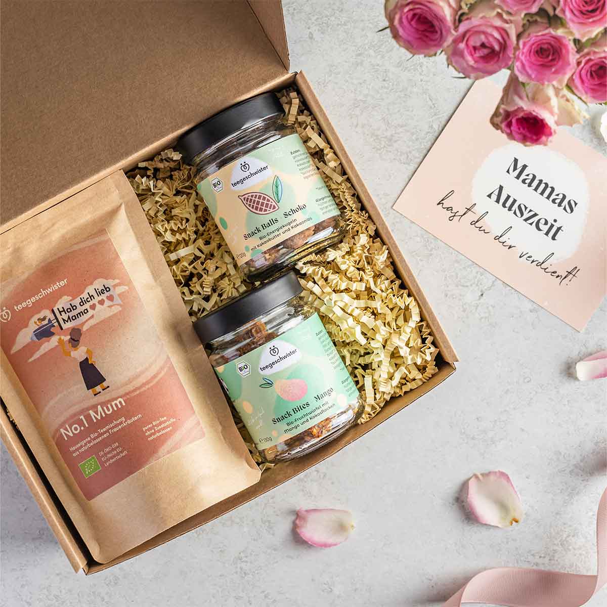 Auf diesem Bild sieht man die offene Geschenkbox mit dem Mama Tee, zwei veganen Snacks und einer Geschenkkarte.