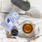 Das Bild zeigt den Gute Nacht Tee auf einer Bettdecke mit einem Buch und aufgegossenem Tee.