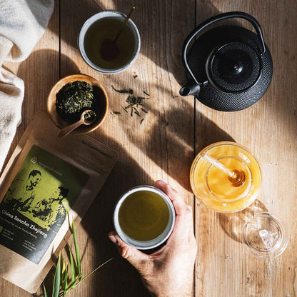 Auf diesem Bild sieht man die Verpackung des China Sencha mit losem Tee und einen Aufguss in einer Teekanne.