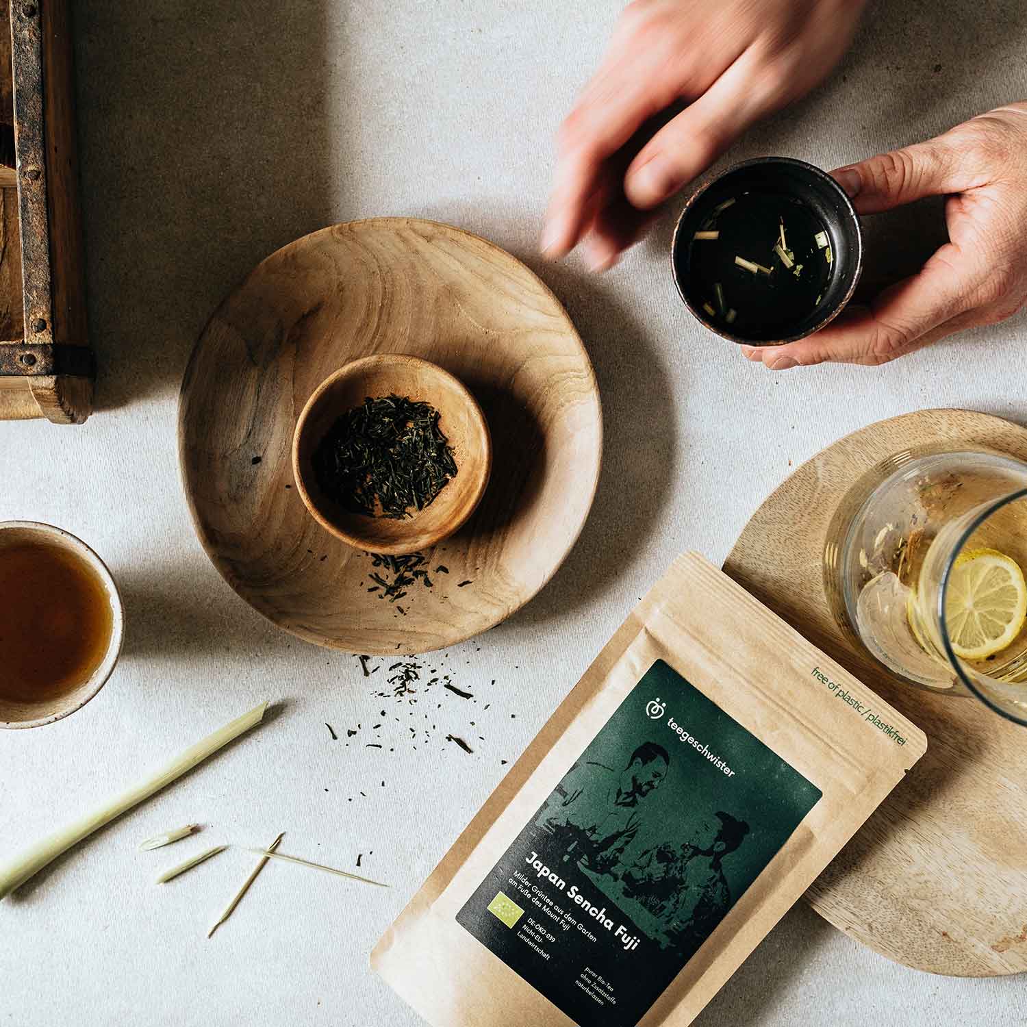 In diesem Bild sieht man die Tee Verpackung des Japan Sencha und den losen Tee in einer Schale. Eine Hand hält einen aufgegossenen Sencha Tee.